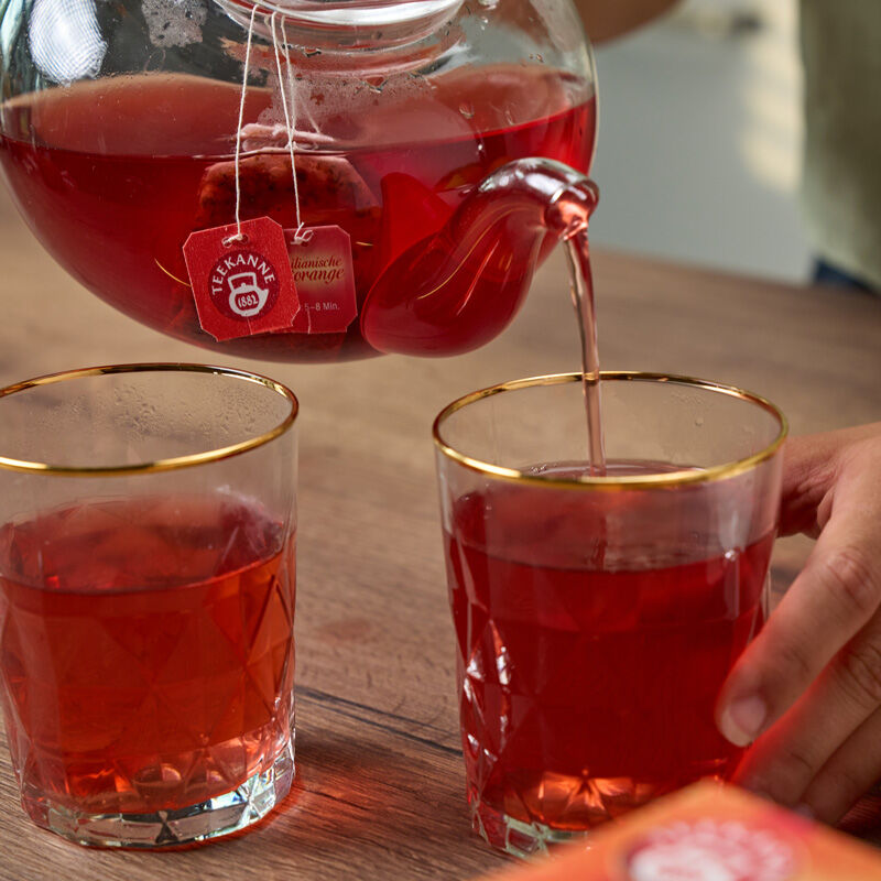 Eine Nahaufnahme von Ländertee Mix & Match Tee, der aus einer Glasteekanne in ein goldumrandetes Glas gegossen wird, neben einem weiteren auf einem Holztisch. Eine Hand hält eines der Gläser und fängt den perfekten Teemoment ein.