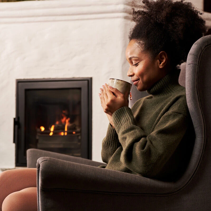 Eine Frau in einem grünen Rollkragenpullover sitzt in einem grauen Sessel vor einem brennenden Kamin und lächelt sanft, während sie mit beiden Händen eine Tasse Bio-Wintertee Mix & Match hält.