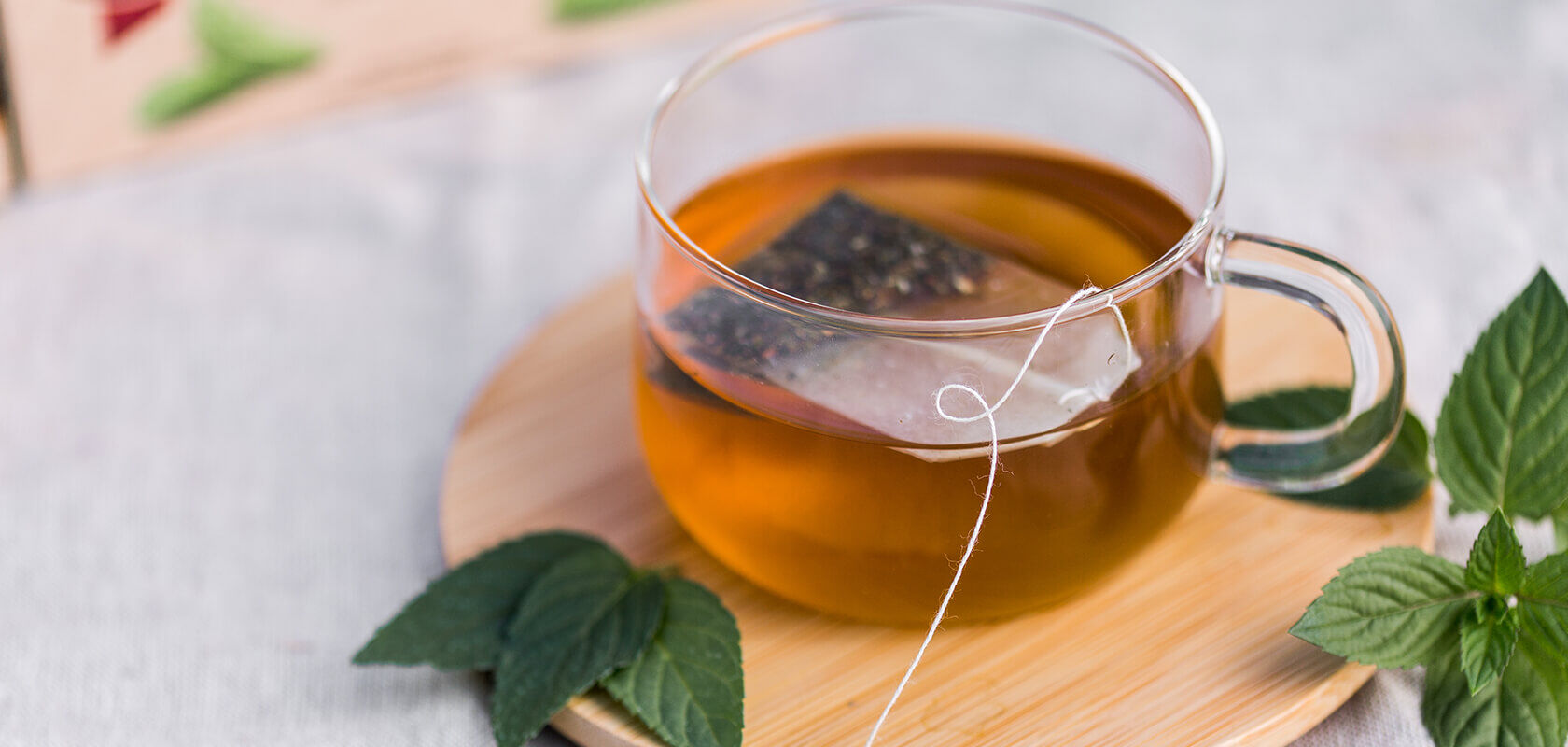 Glas mit schwarzem Tee und Teebeutel auf rundem Holzbrett, umgeben von frischen grünen Blättern