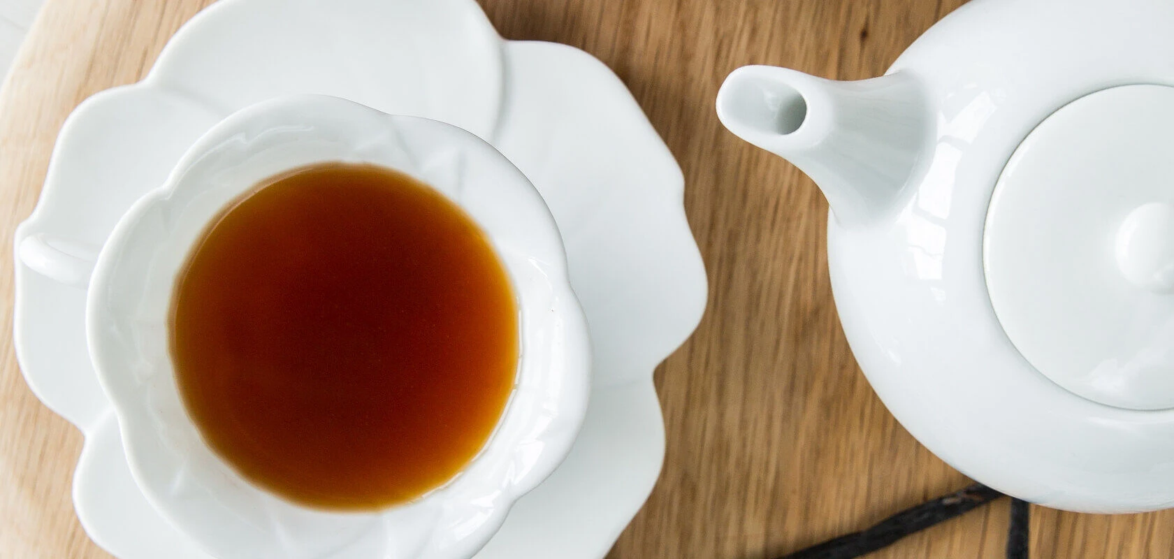 Eine weiße Teetasse mit Teefix (Familienpackung) Schwarztee steht auf einer passenden Untertasse neben einer weißen Teekanne, alles auf einer Holzoberfläche. Genießen Sie dieses exquisite Naturprodukt und genießen Sie die reichen Aromen Indiens an Ihrem T