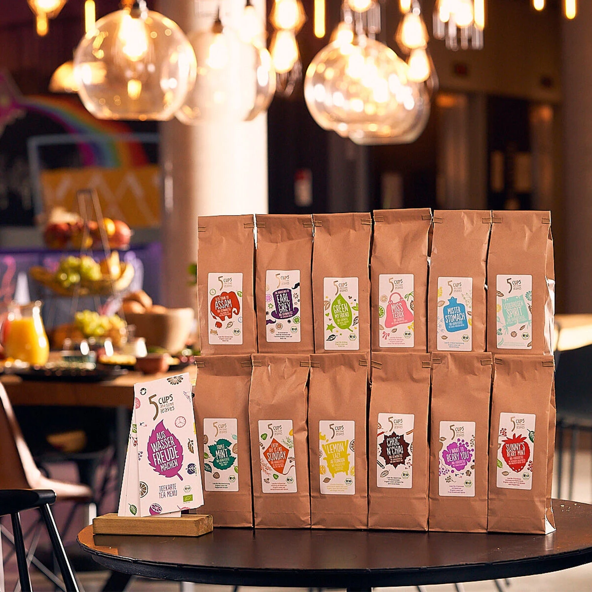Zwölf braune Papiertüten, darunter das 5 CUPS I Want to Berry You Bio Loser Tee, stehen aufrecht in zwei Reihen auf einem Tisch, davor eine Tüte und eine kleine Schachtel. Die Kulisse bildet ein gemütliches Café mit warmer Beleuchtung.