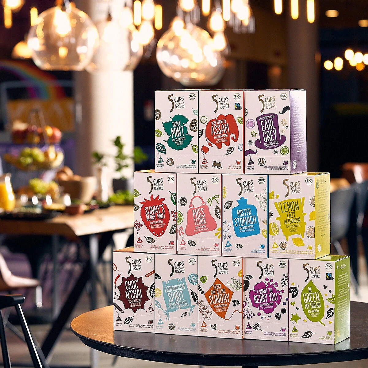 Eine Ausstellung farbenfroher 5 CUPS AND SOME SUGAR-Teeboxen, darunter der 5 CUPS Lederhosen Spirit Bio Pyramidenbeutel, in drei Etagen auf einem Tisch angeordnet, mit einem verschwommenen Café-Interieur und Hängelampen im Hintergrund.