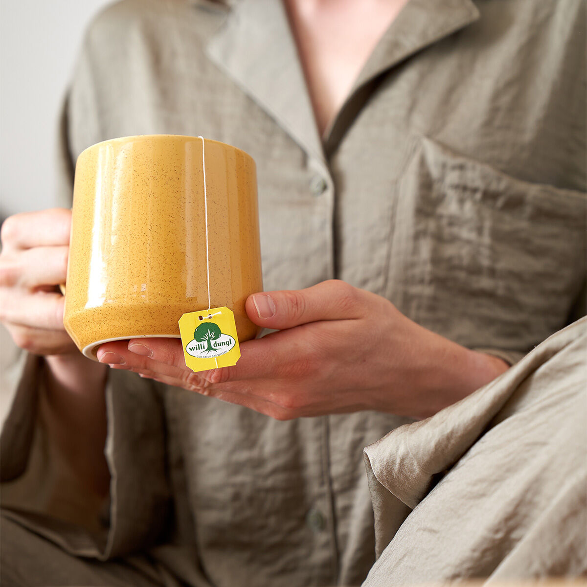 Eine Person im hellbraunen Pyjama hält eine große gelbe Tasse Tee mit einem sichtbaren gelben Etikett mit der Aufschrift „Wach & Konzentriert“ auf der Teebeutelschnur.