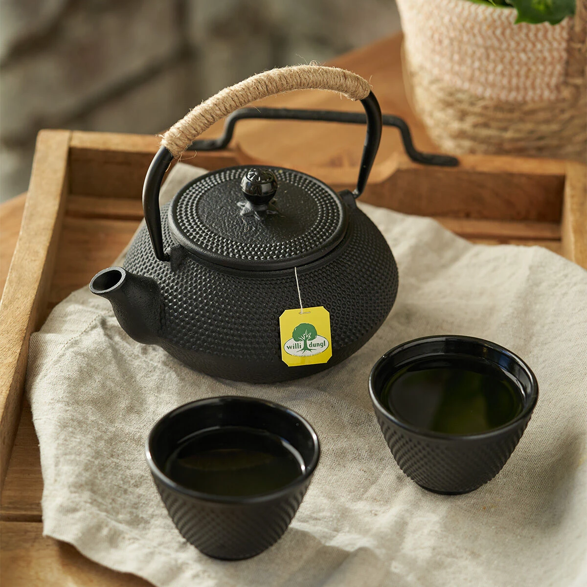 Eine schwarze Teekanne aus Gusseisen mit garnumwickeltem Henkel steht auf einem beigen Tischtuch neben zwei Teetassen mit Antioxidativ Grüntee BIO, die auf einem Holztablett angeordnet sind. An der Teekanne ist ein grünes Teebeuteletikett für Antioxidativ
