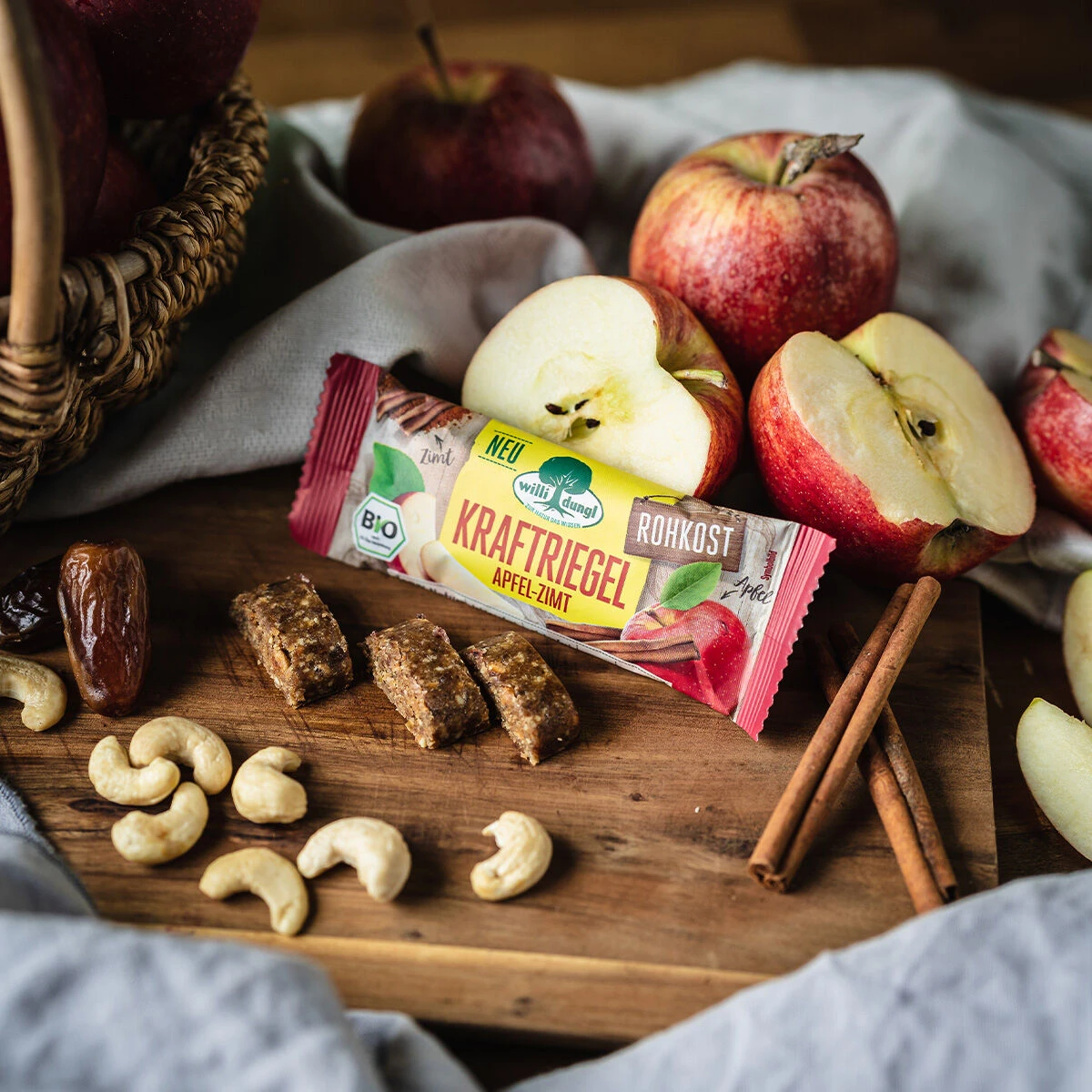 Ein Kraftriegel Apfel-Zimt Bio-Snack liegt auf einem Holzbrett neben geschnittenen Äpfeln, Cashewnüssen, Datteln und Zimtstangen. Ein Korb mit Äpfeln im Hintergrund trägt zur gemütlichen, rustikalen Atmosphäre bei.
