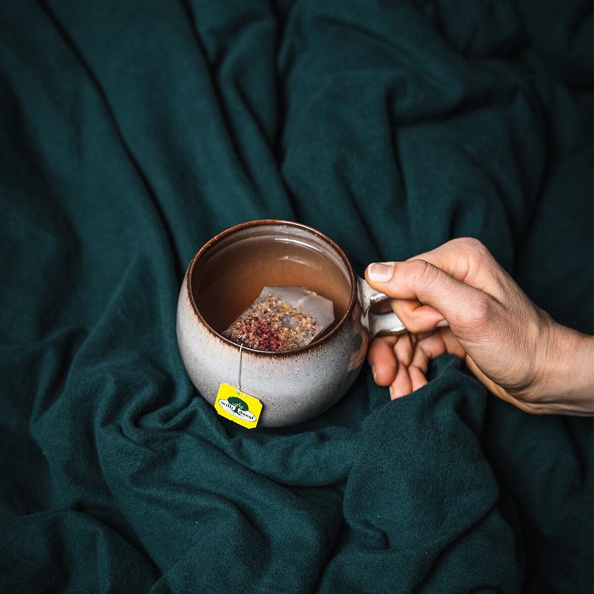 Eine Hand hält eine Keramiktasse mit einem Teebeutel darin, die auf einer dunkelgrünen, weichen Decke liegt. Das gelbe Etikett des Teebeutels trägt die Aufschrift „Atemfrei BIO“ und verstärkt die gemütliche und ruhige Atmosphäre der Szene.