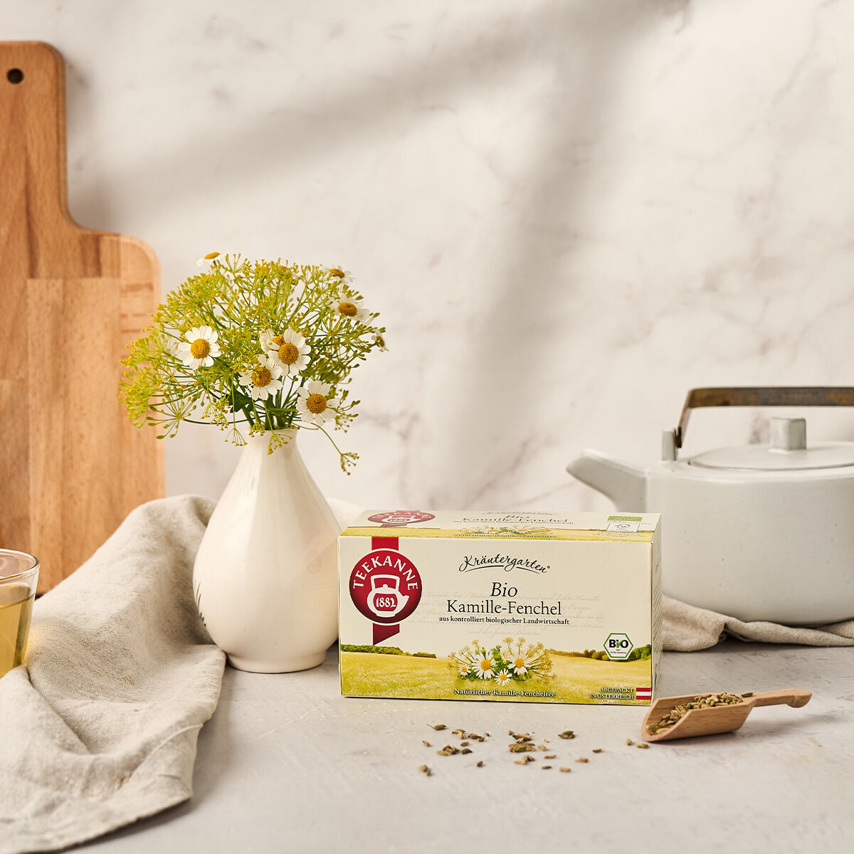 Neben einer weißen Teekanne steht eine Schachtel BIO Kamille-Fenchel, Kamillenblüten, ein Glas Tee und lose Blätter – purer Kräutergenuss aus dem Garten, ganz natürlich und frei von zugesetzten Aromen.