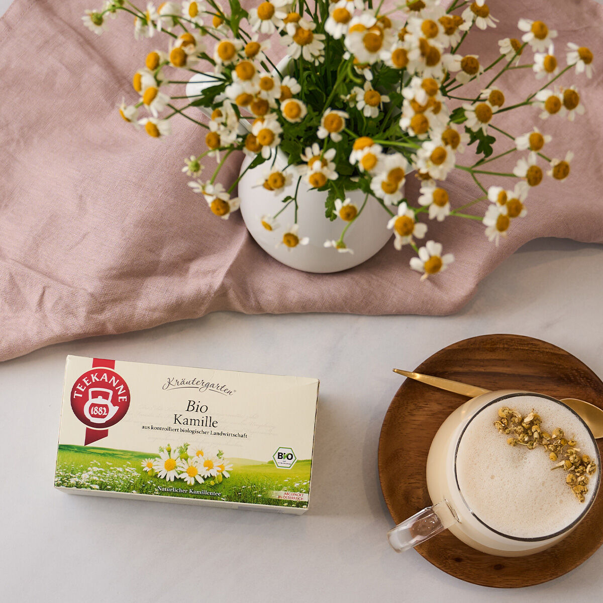 Eine Schachtel BIO Kamille steht auf einem Tisch neben einer Vase mit Kamillenblüten und einer Tasse Tee mit Schaum und Kamille, serviert auf einer Holzuntertasse. Im Hintergrund ist teilweise ein rosa Tuch zu sehen.