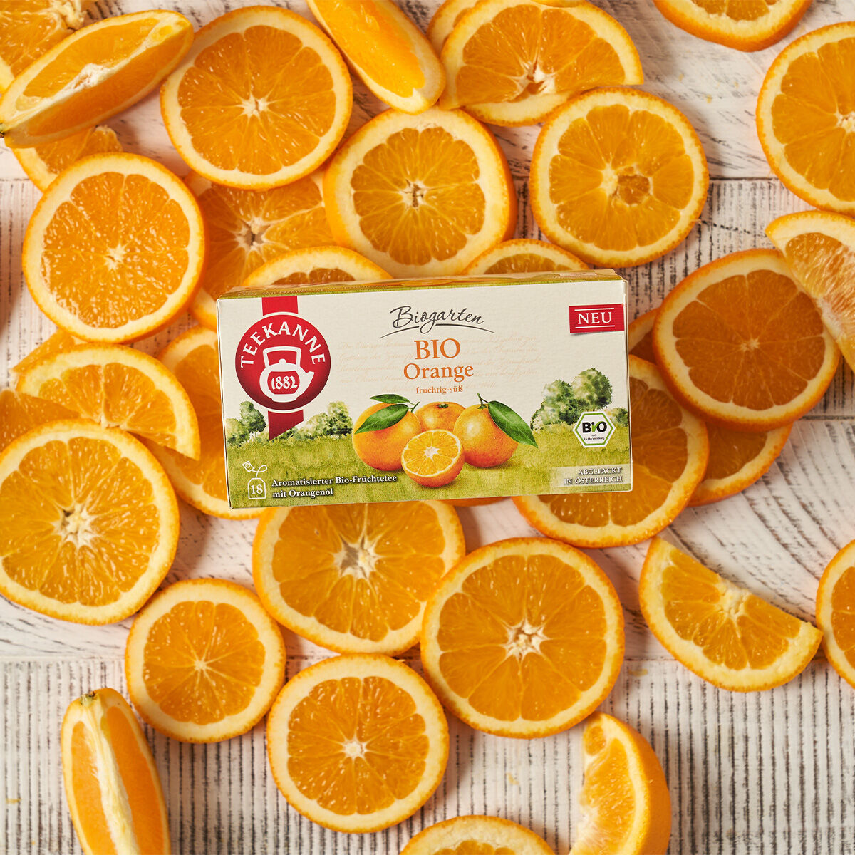 Eine Schachtel BIO Orange Tee steht auf einer weißen Holzoberfläche, umgeben von frischen Orangenscheiben. Die Verpackung zeigt lebendige Orangen- und Blattmotive, wodurch BIO Orange mit seinem auffälligen Design hervorsticht.