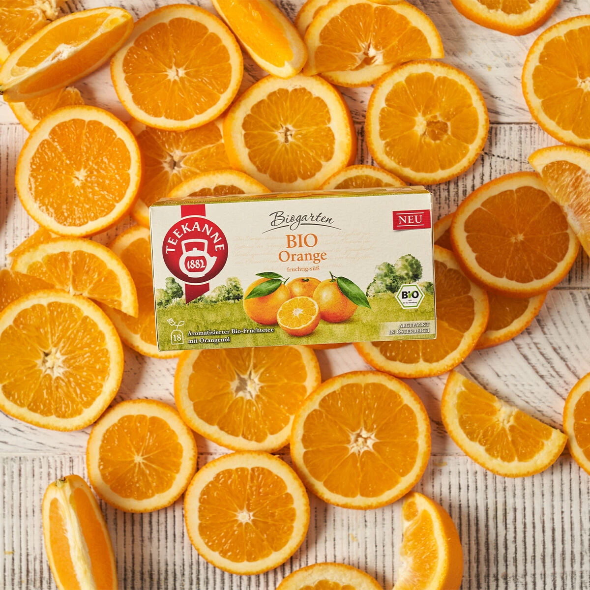 Eine Schachtel BIO Orange Tee steht auf einer weißen Holzoberfläche, umgeben von frischen Orangenscheiben. Die Verpackung zeigt lebendige Orangen- und Blattmotive, wodurch BIO Orange mit seinem auffälligen Design hervorsticht.