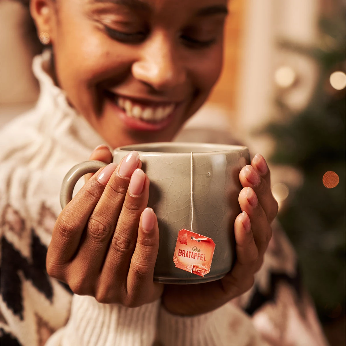 Eine lächelnde Person in einem kuscheligen Pullover hält mit beiden Händen eine graue Tasse Tee mit der Aufschrift „Bio Bratapfel“. An der Tasse hängt ein rotes Etikett, im Hintergrund leuchtet sanft ein verschwommener Weihnachtsbaum.