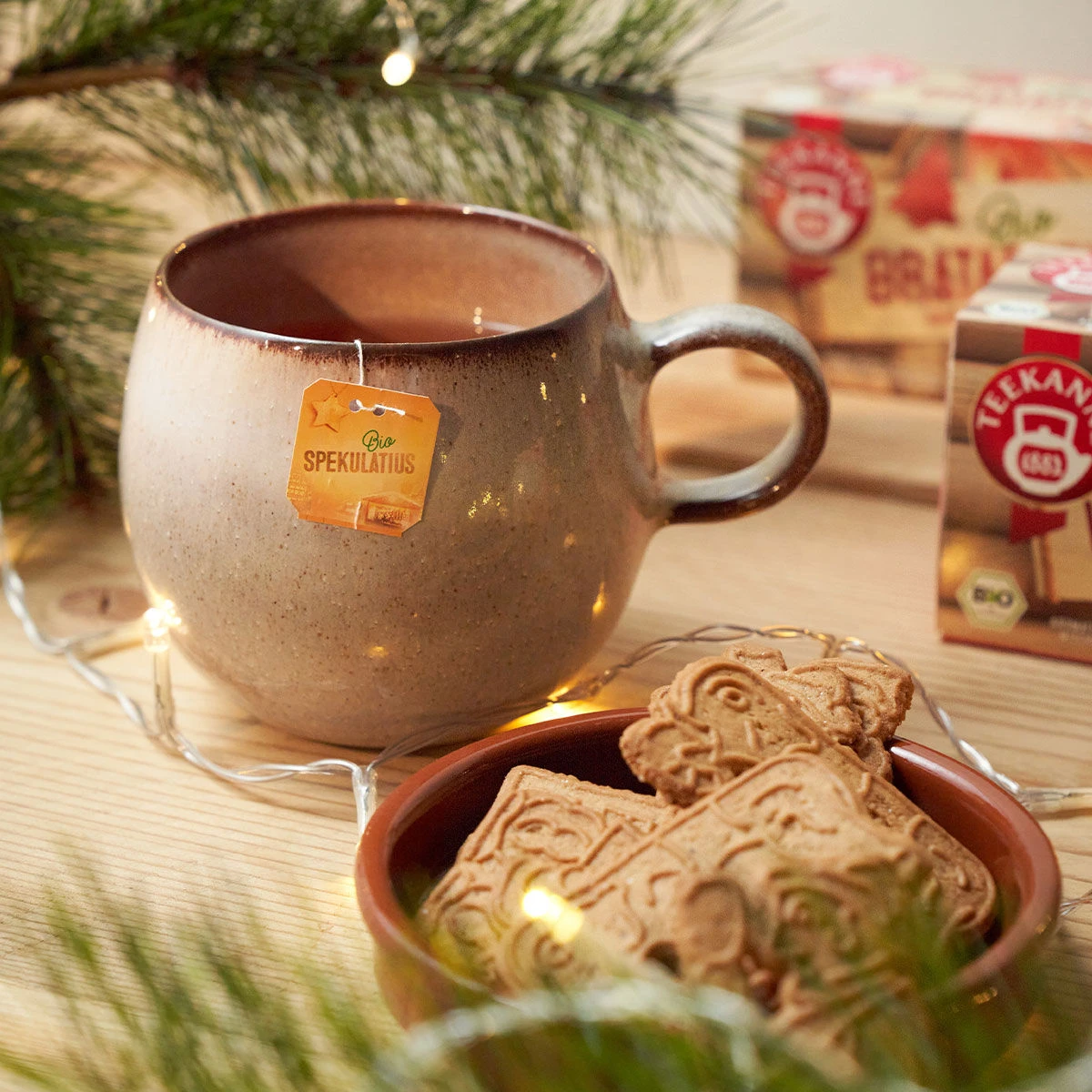 Eine beige Keramiktasse Tee mit dem Etikett eines Bio-Spekulatius-Teebeutels steht auf einem Holztisch neben einer Schale mit Spekulatius-Keksen. Tannenzweige und Weihnachtsbeleuchtung sorgen für eine festliche Atmosphäre.