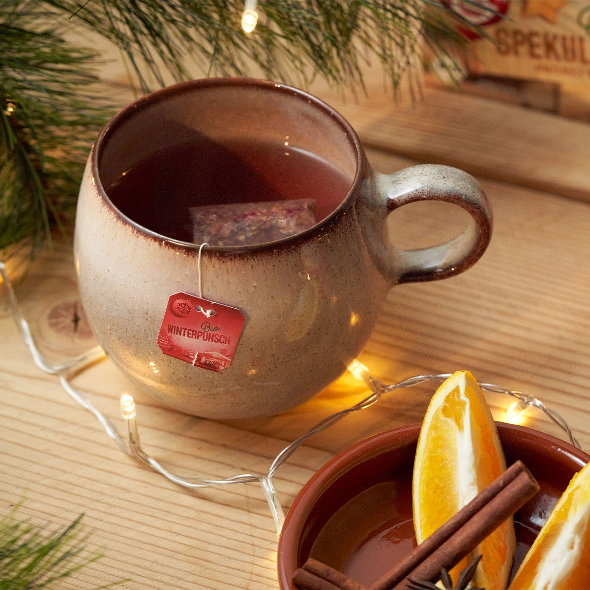 Eine Keramiktasse Tee mit einem roten Bio-Winterpunsch-Teebeutel steht auf einem Holztisch neben festlichen Lichterketten, Tannenzweigen und einer Schale mit Orangenscheiben und Zimtstangen.