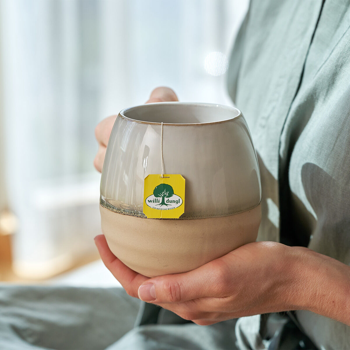 Eine Person in einem hellgrünen Gewand hält einen großen Keramikbecher, an dessen Rand ein gelber Blasen-Nieren Tee BIO-Beutel hängt. Im Hintergrund leuchtet sanftes Sonnenlicht.