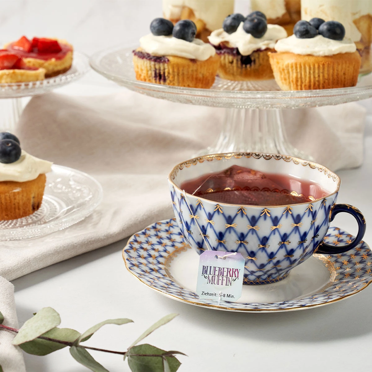 Eine gemusterte Teetasse mit Teefüllung steht auf einer Untertasse neben einem Blueberry-Muffin-Teebeutel. Im Hintergrund sind Cupcakes mit Zuckerguss und Blaubeeren auf Glasständern ausgestellt.