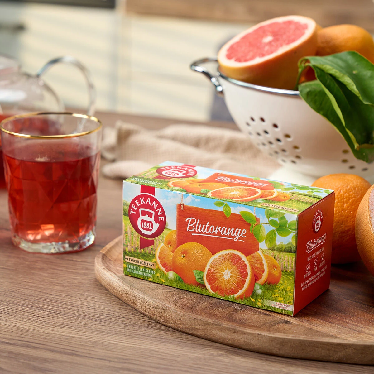Eine Schachtel Blutorange-Tee steht auf einem Holztisch neben einem Glas roten Tee, ganzen Orangen, einer halben Grapefruit und einem weißen Sieb voller bunter Früchte und grüner Blätter – wie ein echter Obstgarten.