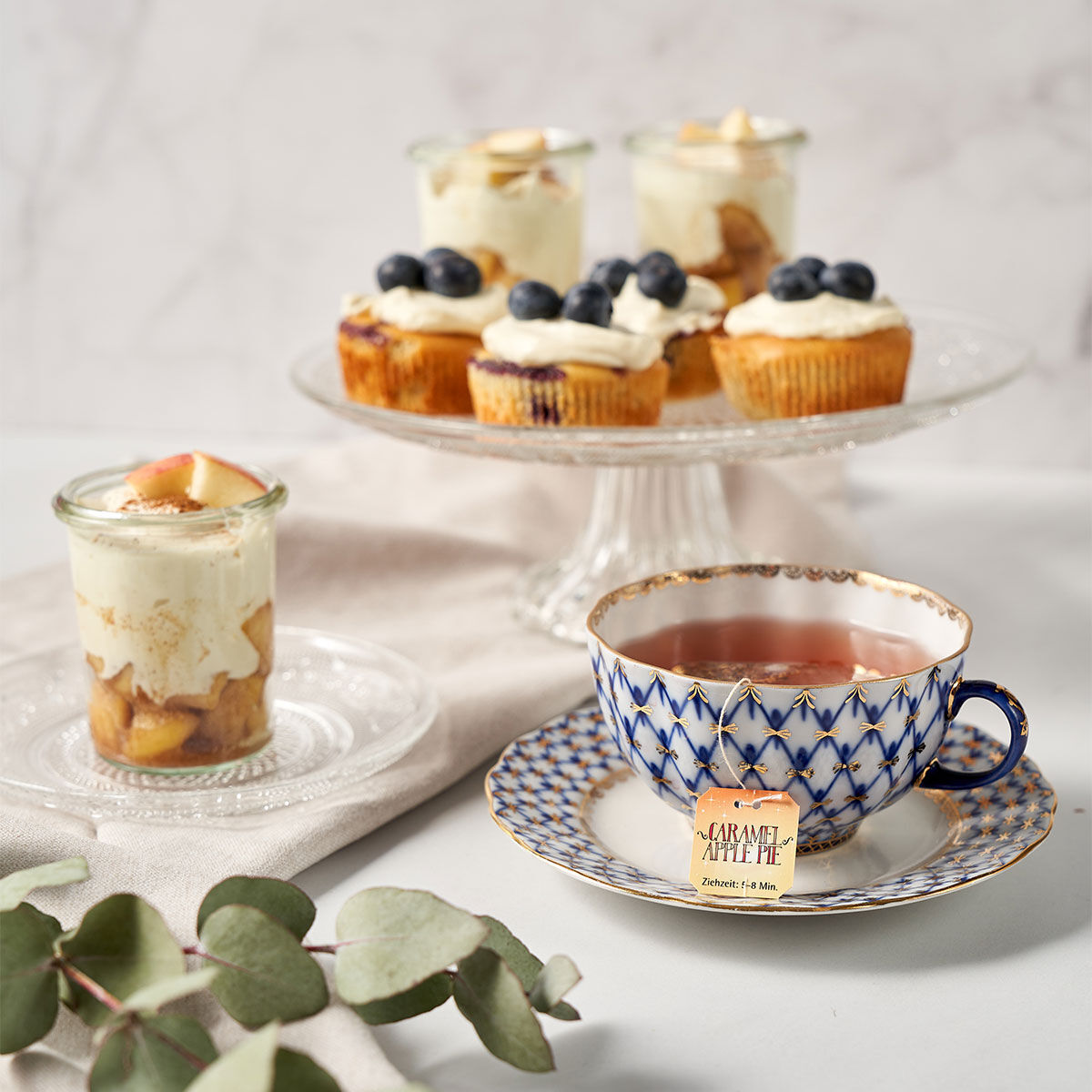 Eine gemusterte Teetasse mit Sweeteas steht auf einer Untertasse neben einem Dessert in einem Glasgefäß. Im Hintergrund ruhen Cupcakes mit Sahne und Blaubeeren auf einem Glasständer, während Eukalyptusblätter und Stücke von Karamell-Apfelkuchen die Szene 
