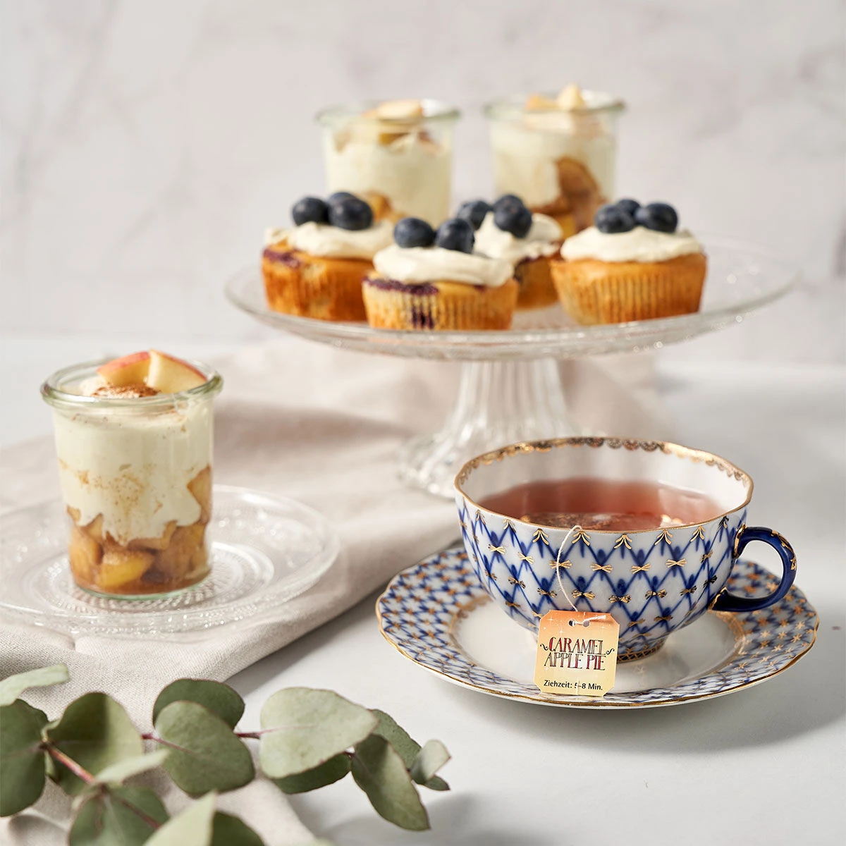 Eine gemusterte Teetasse mit Sweeteas steht auf einer Untertasse neben einem Dessert in einem Glasgefäß. Im Hintergrund ruhen Cupcakes mit Sahne und Blaubeeren auf einem Glasständer, während Eukalyptusblätter und Stücke von Karamell-Apfelkuchen die Szene 