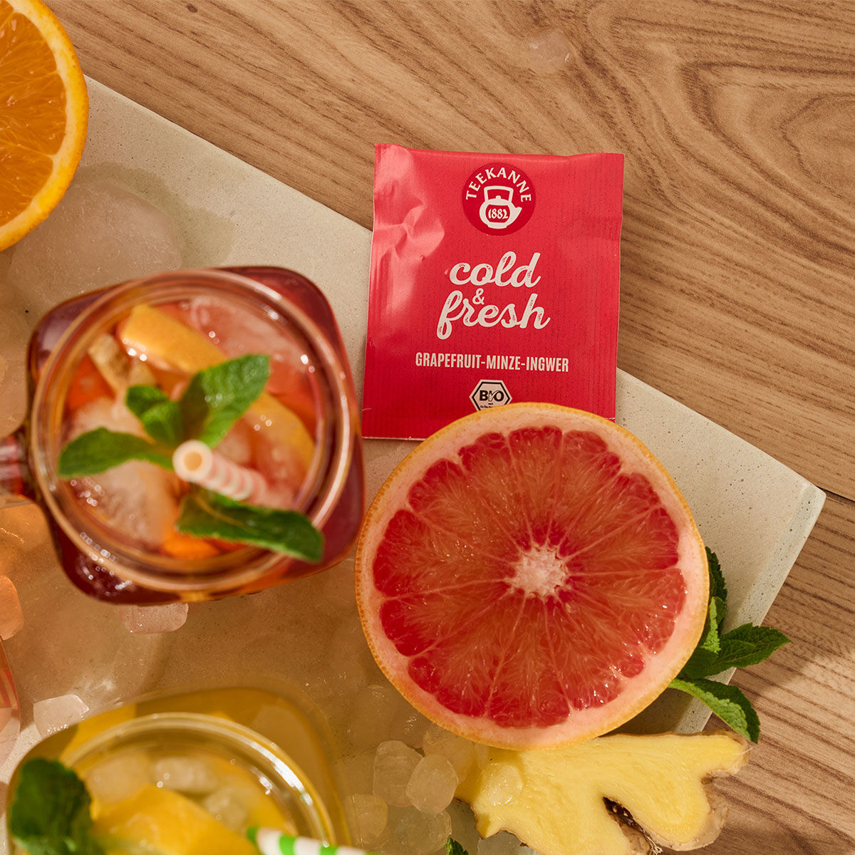 Eine Packung kalter und frischer Grapefruit-Minze-Ingwer Früchtetee steht neben einer halben Grapefruit, einem Glas Eistee mit gestreiftem Strohhalm, Eiswürfeln, Orangenscheiben und Minzblättern – eine perfekte Erfrischung auf dem Holztisch.