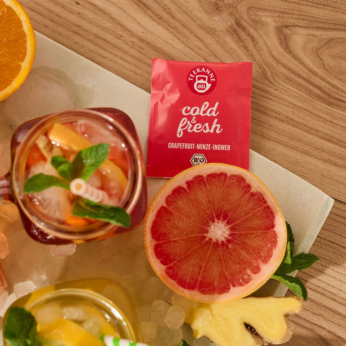Eine Packung kalter und frischer Grapefruit-Minze-Ingwer Früchtetee steht neben einer halben Grapefruit, einem Glas Eistee mit gestreiftem Strohhalm, Eiswürfeln, Orangenscheiben und Minzblättern – eine perfekte Erfrischung auf dem Holztisch.
