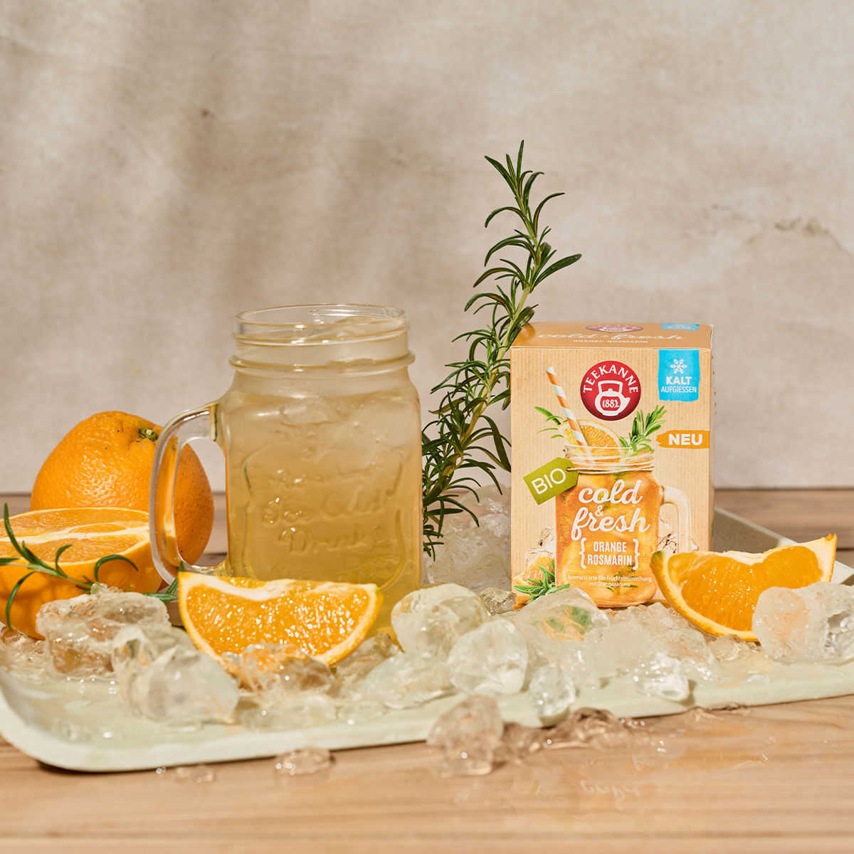 Ein Einmachglas mit eisgekühltem Orangensaft, garniert mit Orangenscheiben und Rosmarin, steht auf einem Tablett neben einer Schachtel kaltem und frischem Orangen-Rosmarin – perfekt für ein erfrischendes Getränk. Orangen und Rosmarin zieren den Hintergrun