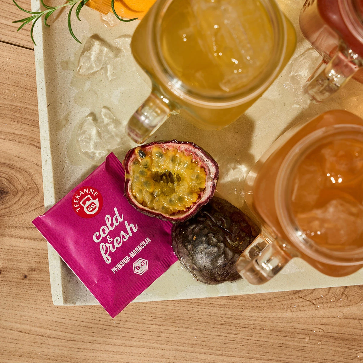 Draufsicht auf ein Tablett mit eisgekühlten Getränken in Einmachgläsern, Eiswürfeln, halbierten und ganzen Passionsfrüchten und einer Packung kalter und frischer Pfirsich-Maracuja auf einer Holzoberfläche – die perfekte Erfrischung für heiße Sommertage.