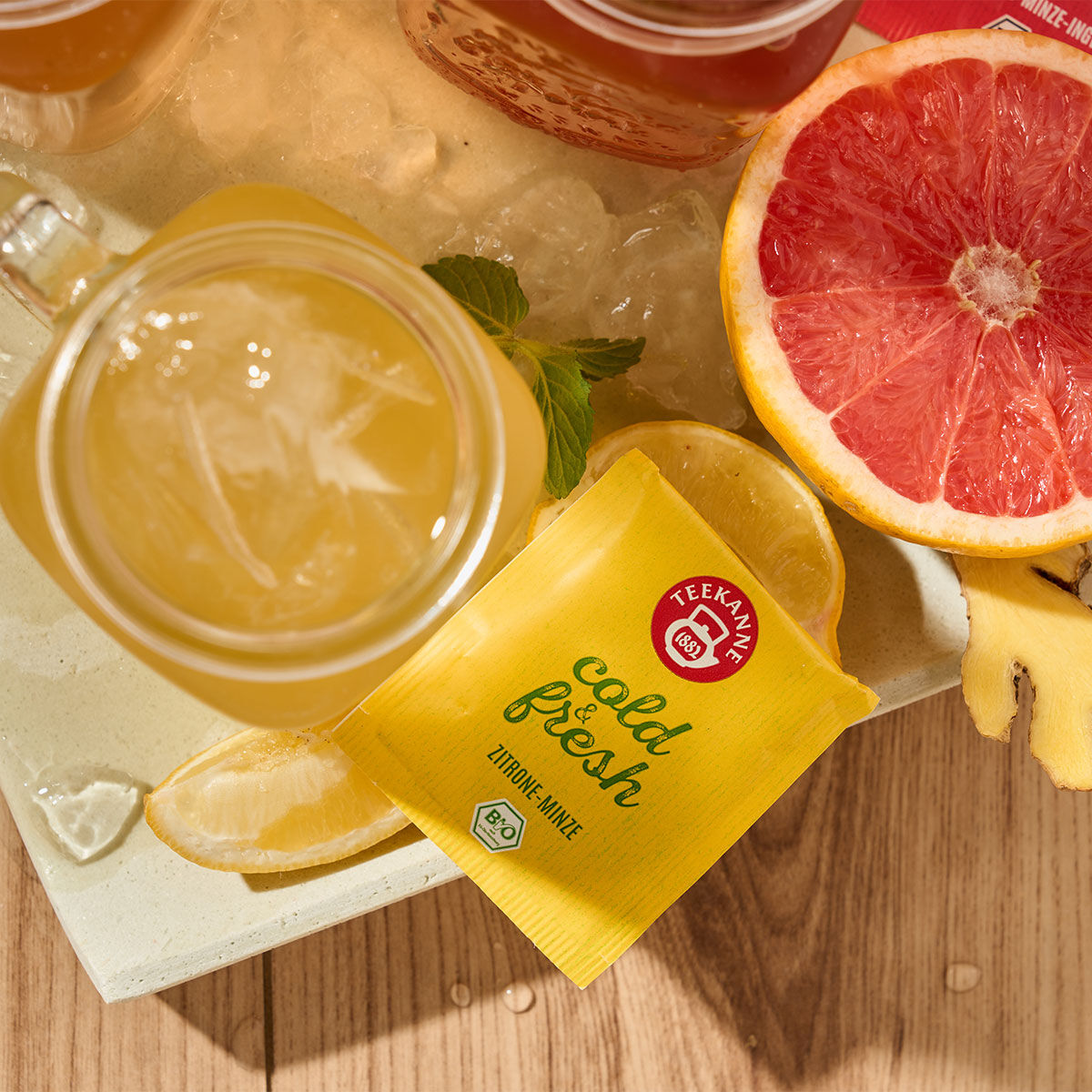 Ein Glas Eistee steht auf einem Tablett mit einer gelben kalten und frischen Zitronen-Minze-Packung, Zitronenscheiben, Ingwer und einer halben Grapefruit auf einer Holzoberfläche – perfekte Erfrischung für heiße Tage.