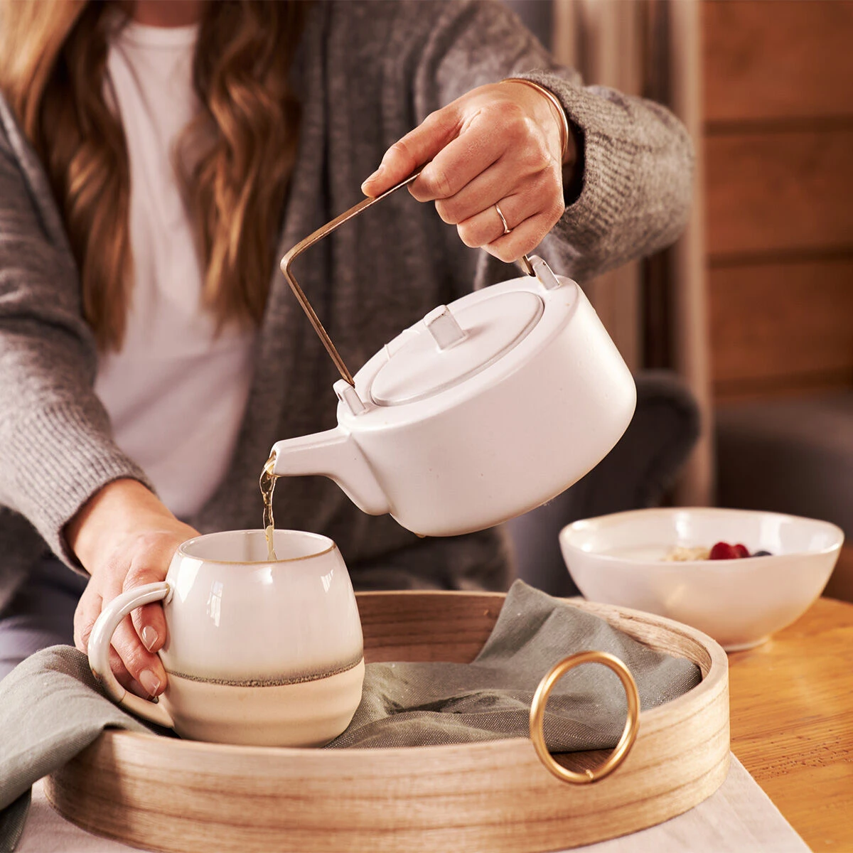 Eine Frau gießt Hochland Grüntee Fairtrade aus einer weißen Teekanne in eine passende Tasse auf einem Holztablett. Neben ihr stehen Haferflocken und Himbeeren. Sie trägt eine graue Strickjacke und sitzt in einem gemütlichen, exklusiven Ambiente.
