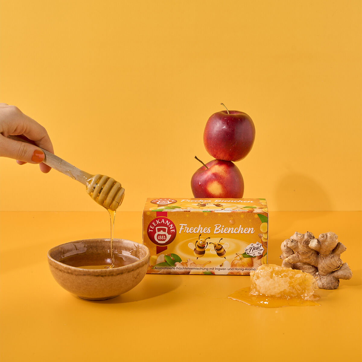 Eine Hand träufelt Honig aus einem Schöpflöffel in eine Schüssel. Daneben liegen Apfelscheiben, Ingwerwurzel, ein Stück Honigwabe und eine Schachtel Freches Bienchen-Tee, alles arrangiert auf einem leuchtend gelben Hintergrund.