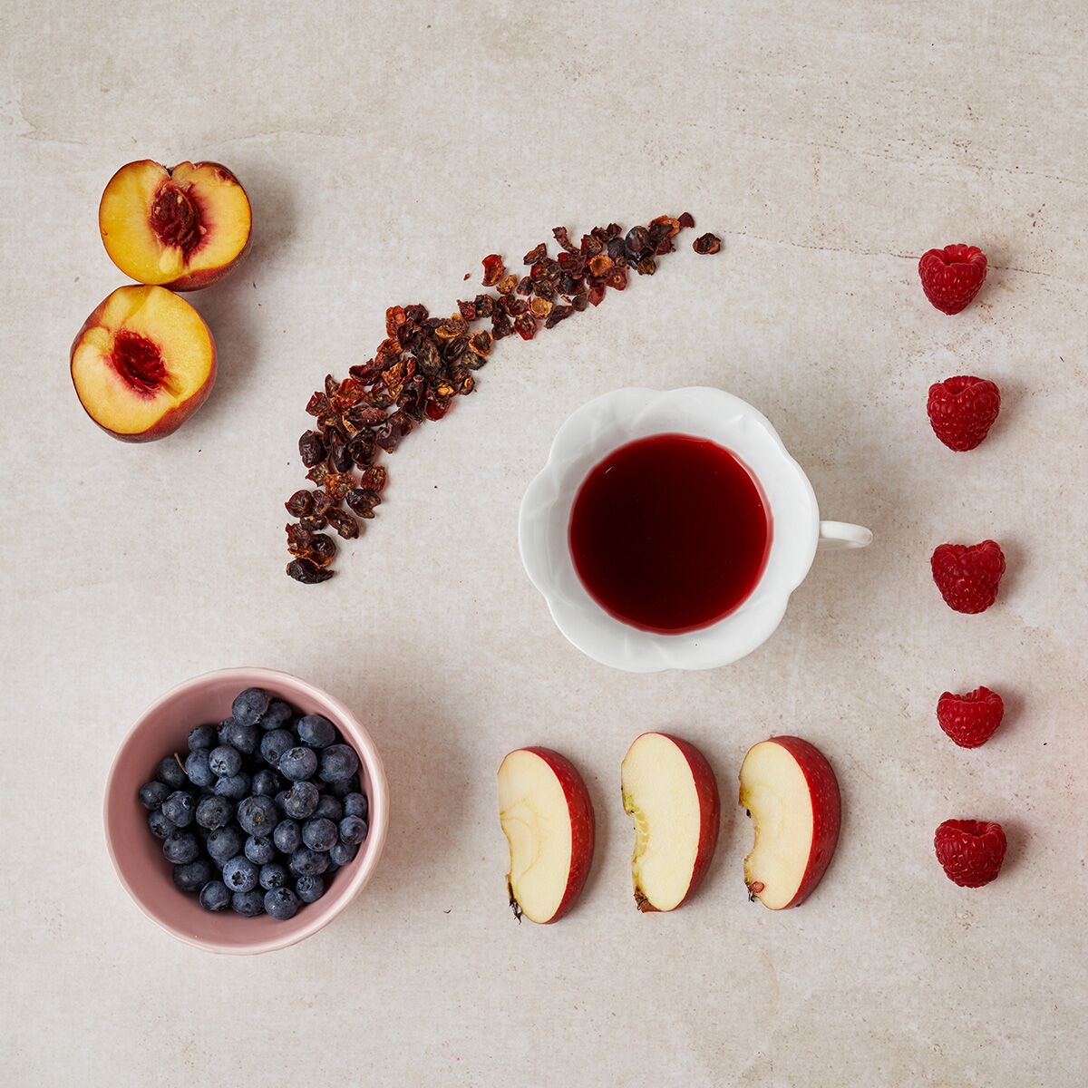 Ein Flatlay zeigt Pfirsichhälften, getrocknete Früchteteeblätter, eine Tasse roten Tee, Himbeeren, Blaubeeren in einer Schüssel und drei Apfelscheiben auf einer hellen Oberfläche.