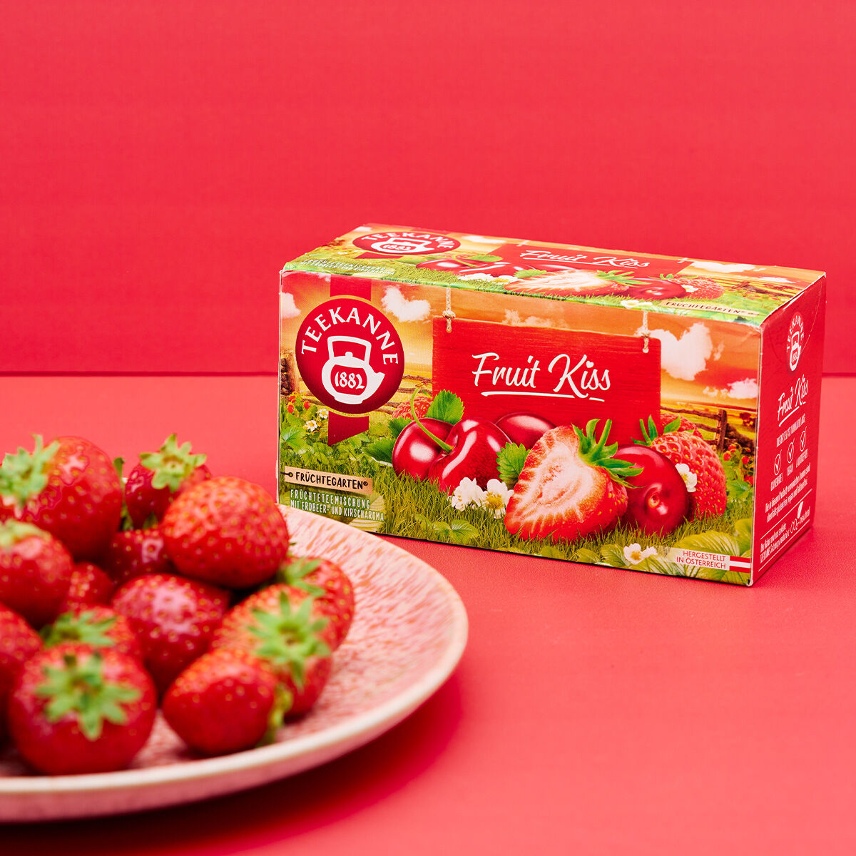 Eine Schachtel Fruit Kiss-Tee mit Erdbeeren und roten Früchten auf der Verpackung steht auf einer roten Fläche neben einem Teller mit frischen Erdbeeren, alles vor einem leuchtend roten Hintergrund.