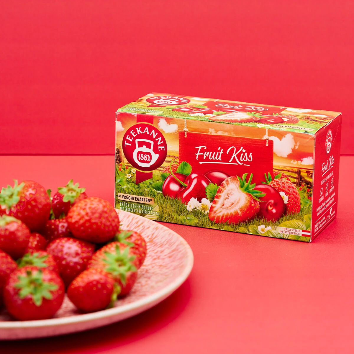 Eine Schachtel Fruit Kiss-Tee mit Erdbeeren und roten Früchten auf der Verpackung steht auf einer roten Fläche neben einem Teller mit frischen Erdbeeren, alles vor einem leuchtend roten Hintergrund.