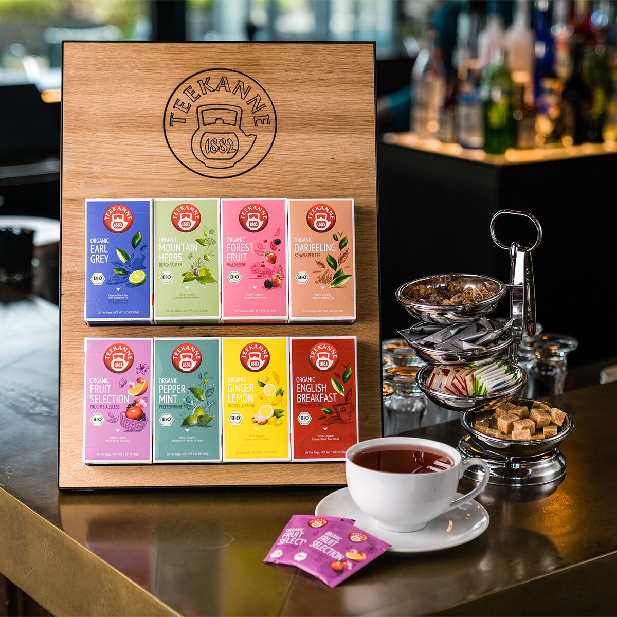Auf einem Holzständer auf der Theke eines Cafés sind Päckchen Bio-Earl-Grey-Tee neben einer Tasse schwarzen Tee, Zuckerwürfeln und Keksen ausgestellt. Im Hintergrund sind die Flaschen unscharf zu sehen.