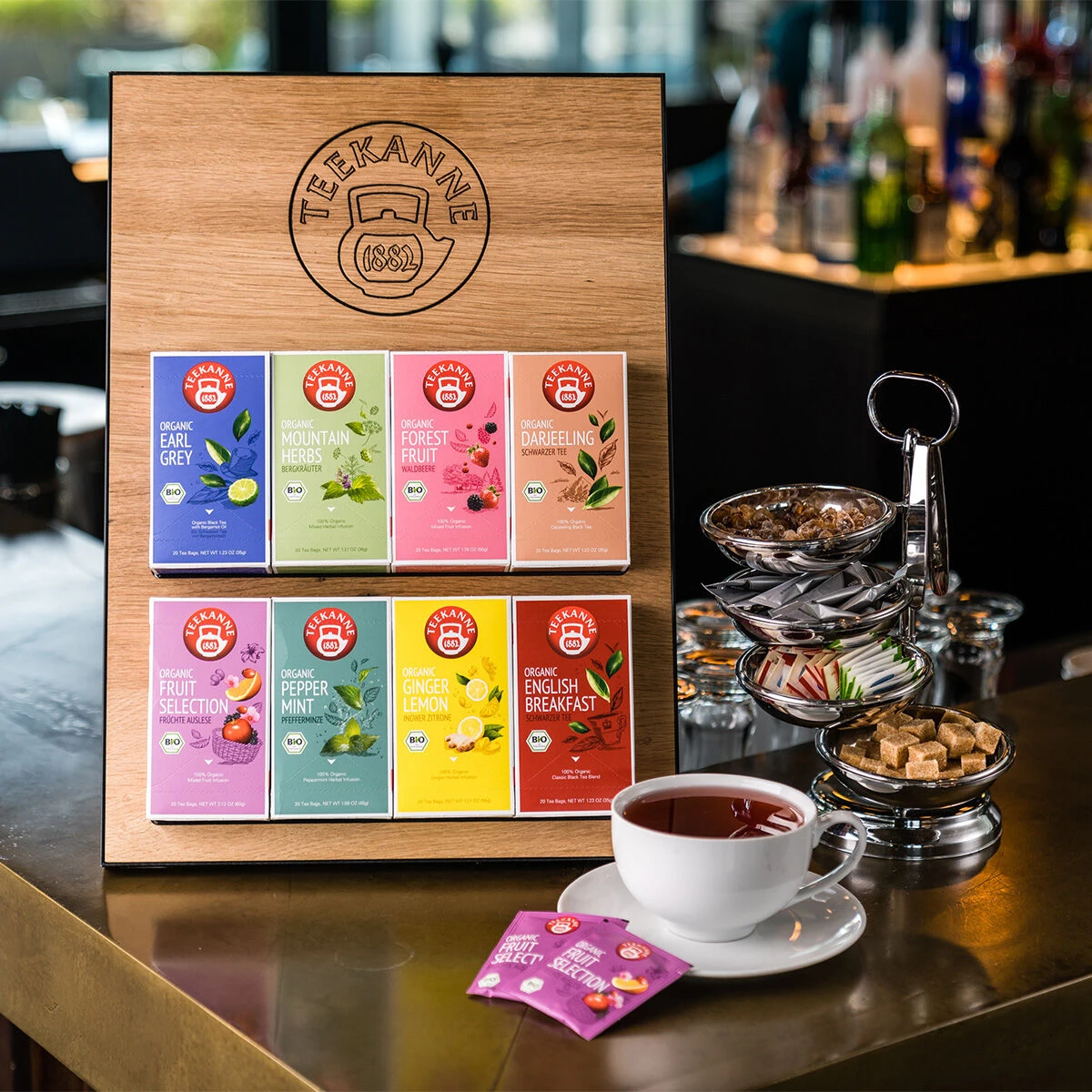 Eine Auswahl verschiedener Teekanne-Teepackungen, darunter Bio-Pfefferminze, steht zusammen mit einer Tasse Tee und Zuckerwürfeln – ideal für Gastronomie oder Büro – auf einem Holzbrett auf einer polierten Theke vor einem unscharfen Bar-Hintergrund.