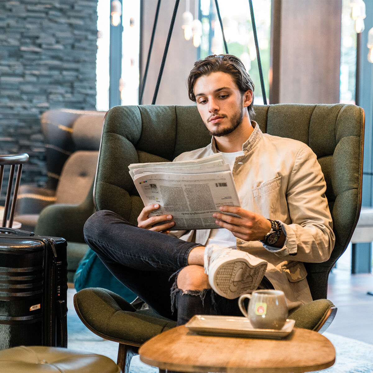 Ein junger Mann entspannt in einem grünen Sessel und liest mit übereinandergeschlagenen Beinen Zeitung. Neben ihm steht ein Koffer, auf dem Tisch eine Tasse 8-Kräuter-Tee. Die moderne Lounge strahlt Komfort und gehobenes Gastronomie-Flair aus.