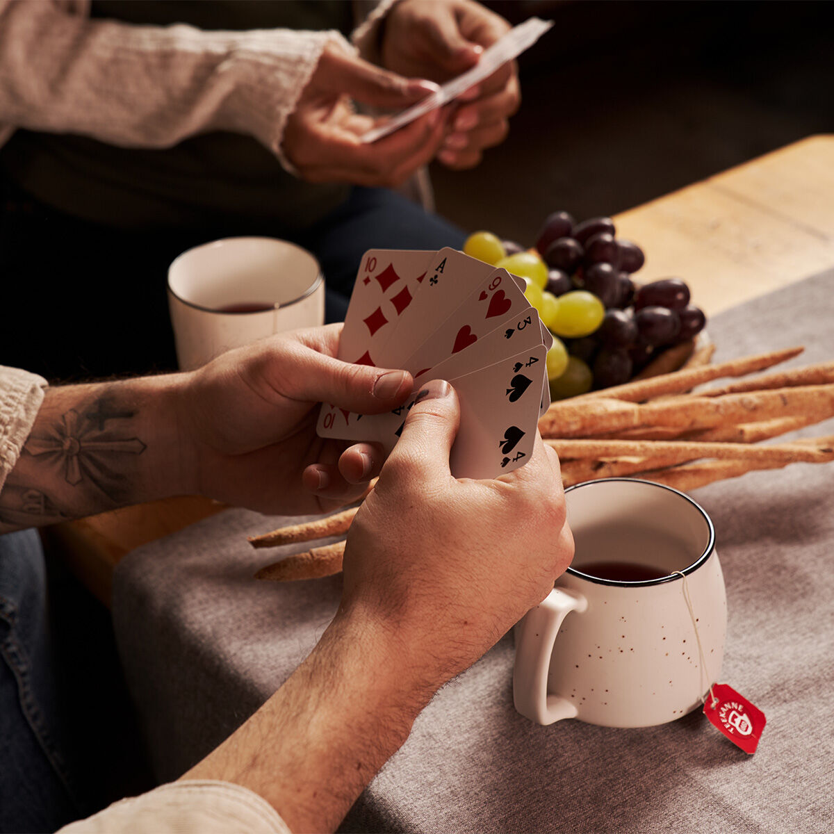 Zwei Personen spielen Karten an einem Tisch mit Grissini, Weintrauben und Glühfix-Bechern. Eine Person hält Spielkarten, während die andere im Hintergrund ihre Karten sortiert.