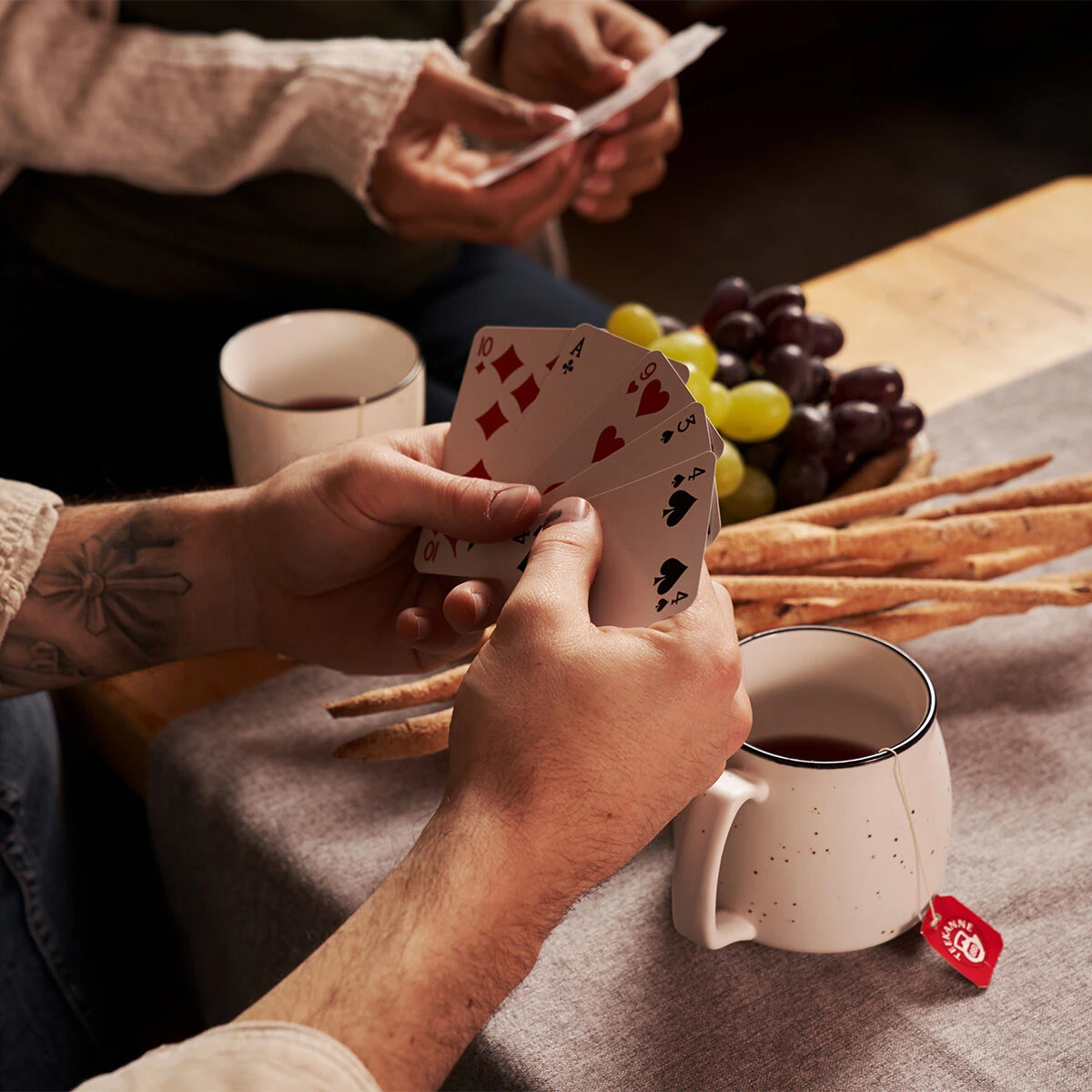 Zwei Personen spielen Karten an einem Tisch mit Grissini, Weintrauben und Glühfix-Bechern. Eine Person hält Spielkarten, während die andere im Hintergrund ihre Karten sortiert.