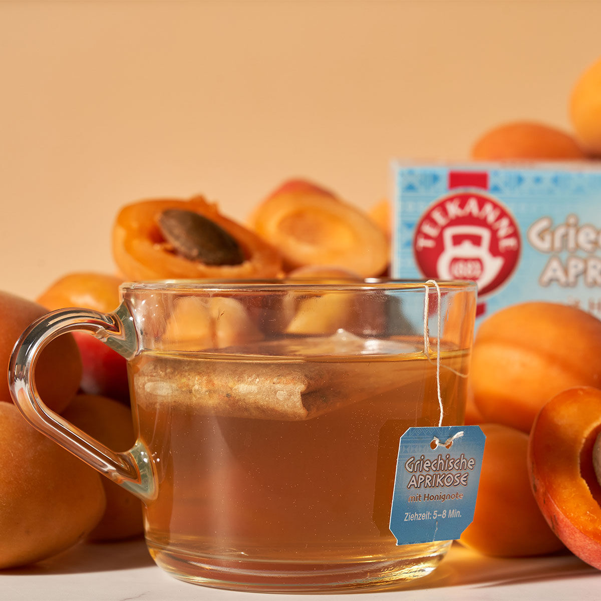 Eine Tasse Griechischer Aprikosentee aus klarem Glas mit einem beschrifteten Teebeutel steht vor frischen Aprikosen und einer Schachtel Griechischer Aprikosen vor einem warmen, hellen Hintergrund.