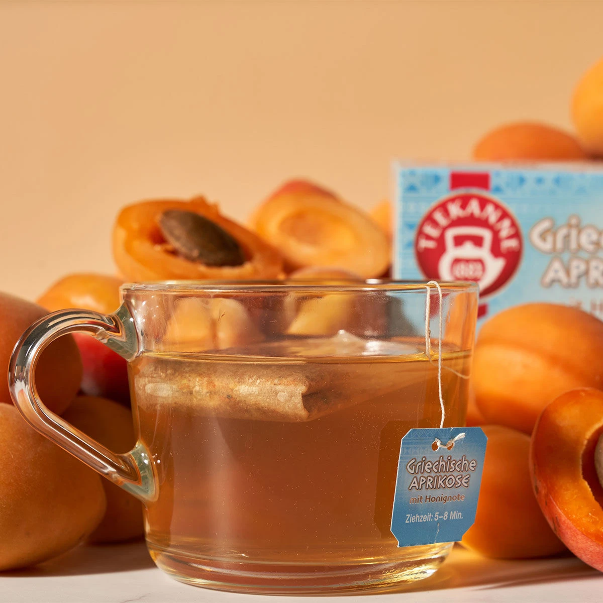Eine Tasse Griechischer Aprikosentee aus klarem Glas mit einem beschrifteten Teebeutel steht vor frischen Aprikosen und einer Schachtel Griechischer Aprikosen vor einem warmen, hellen Hintergrund.
