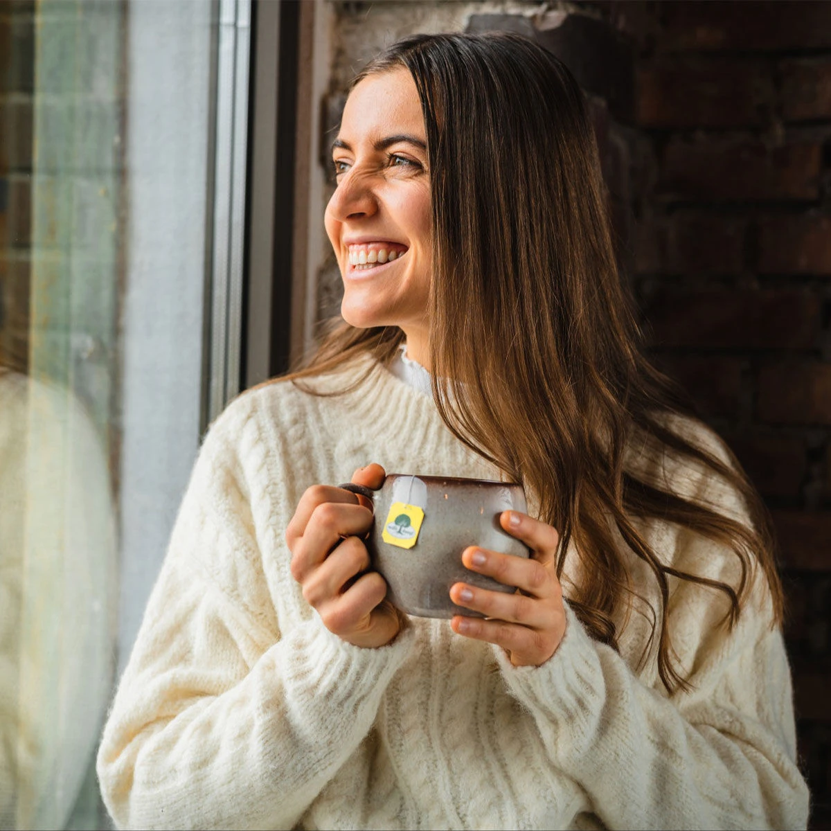 Eine Frau mit langen braunen Haaren in einem weißen Pullover lächelt und hält eine Gute Laune BIO-Tasse am Fenster. Warmes Licht beleuchtet ihr Gesicht, hinter ihr ist eine Backsteinmauer zu sehen.