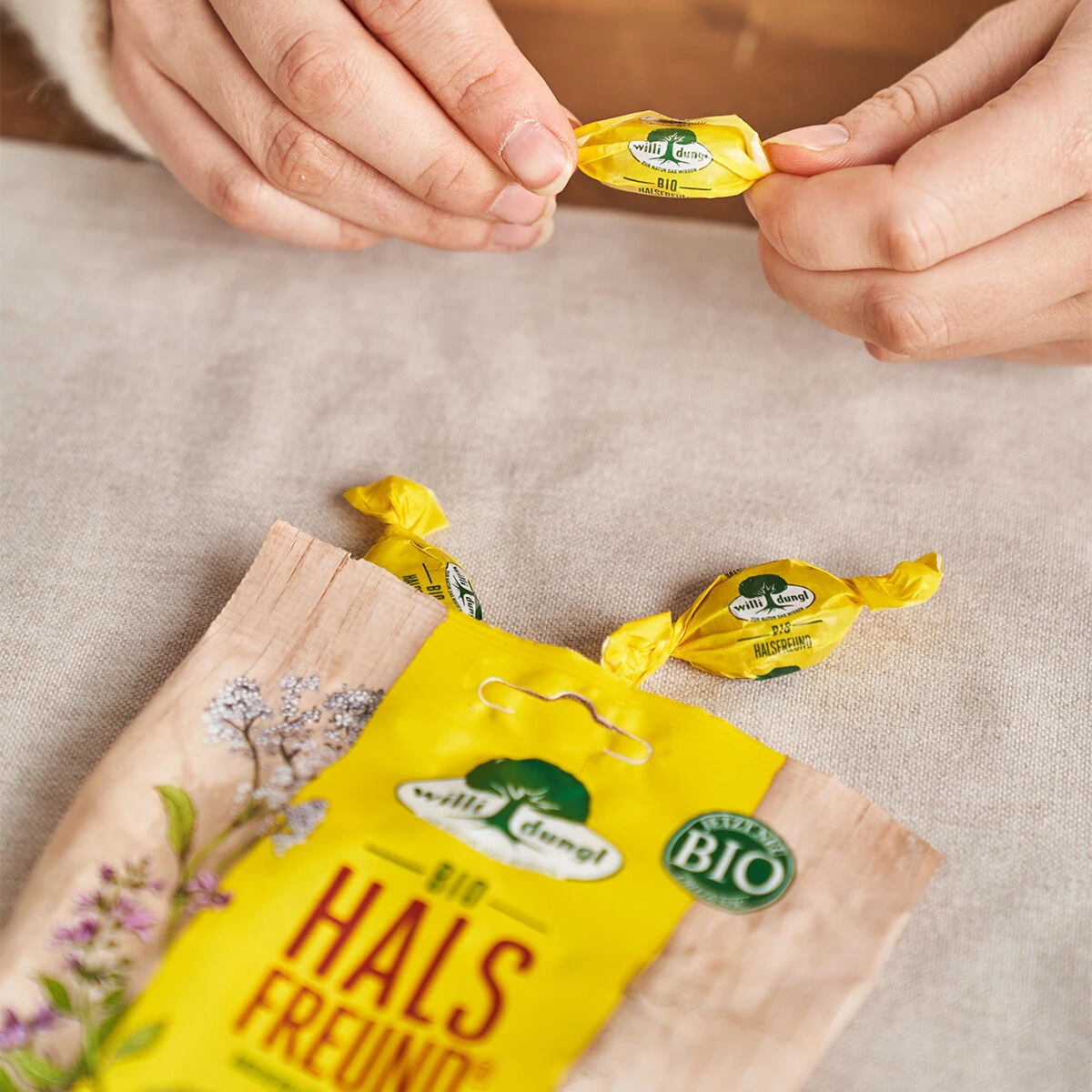 Jemand packt ein gelbes Halsfreund-Bonbon (65 g) aus seiner Verpackung auf einer hellen Stoffoberfläche aus. Neben der offenen Tüte liegen weitere eingewickelte Bonbons.