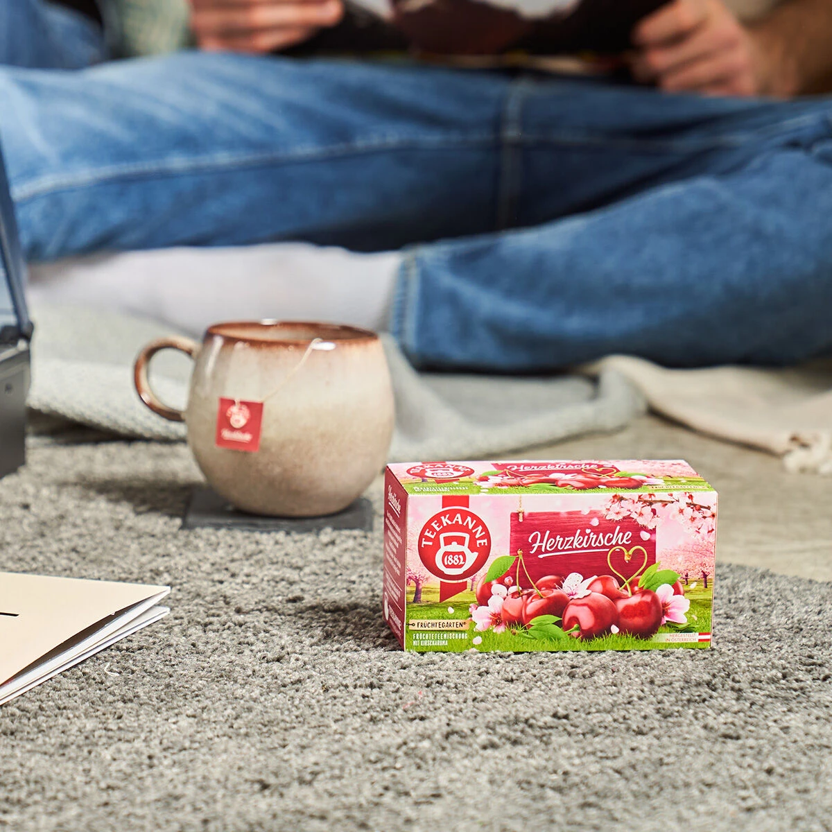 Auf einem grauen Teppich steht eine Schachtel Herzkirsche-Tee neben einer Keramiktasse mit Teebeutel und einem offenen Notizbuch; im Hintergrund sitzt jemand in Jeans.