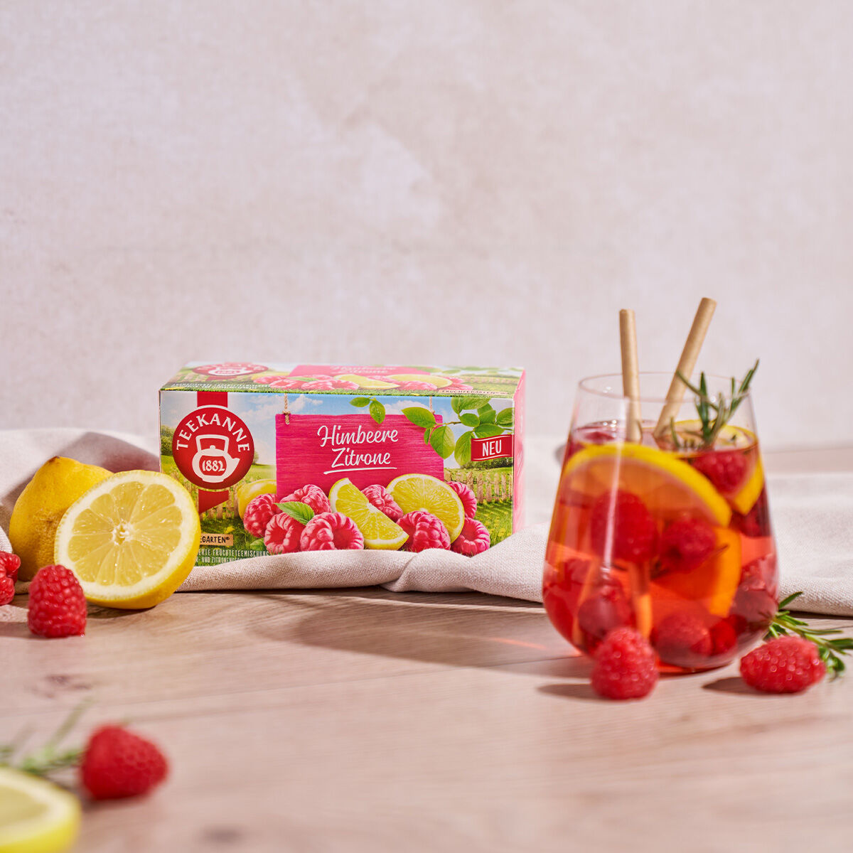 Eine Schachtel Himbeer-Zitronen-Tee steht auf einer Holzoberfläche neben einer Zitronenscheibe, frischen Himbeeren und einem Glas Eistee, dekoriert mit Himbeeren, Zitronenscheiben und Rosmarin.