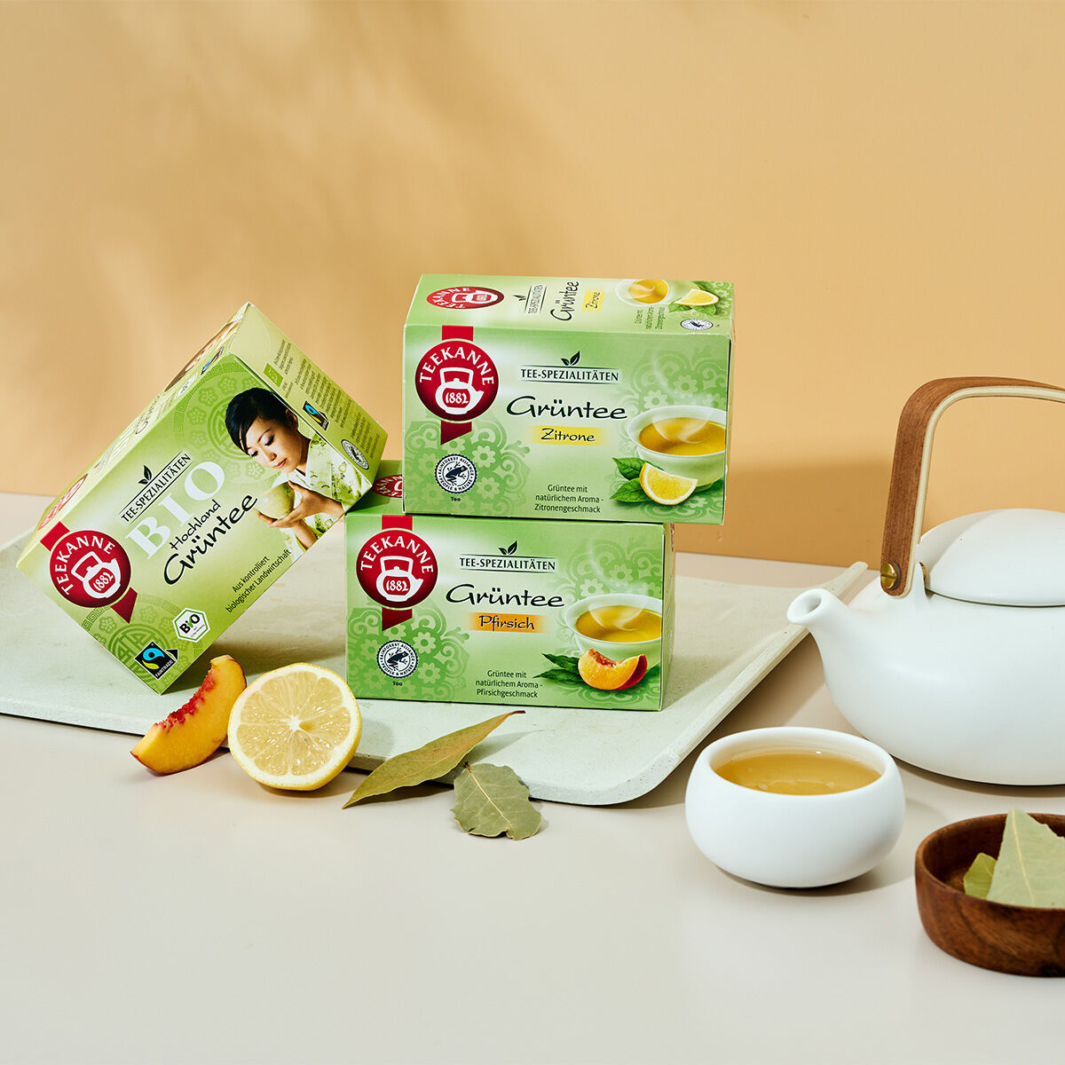 Drei Kartons Hochland Grüntee BIO, Fairtrade & RFA sind mit einer weißen Teekanne, einer gefüllten Teetasse, einer Zitronenscheibe, einem Pfirsich und Teeblättern auf beigem Untergrund arrangiert. Zwei Kartons sind gestapelt; ein weiterer steht daneben un