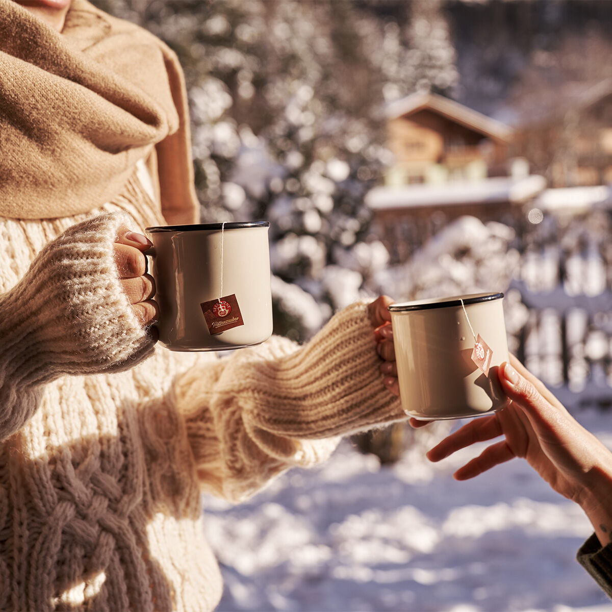 Zwei Menschen in kuscheligen Pullovern und Schals halten draußen im Schnee Metallbecher mit Hüttenzauber in der Hand, im Hintergrund sind Bäume und eine Holzhütte sanft verwischt.