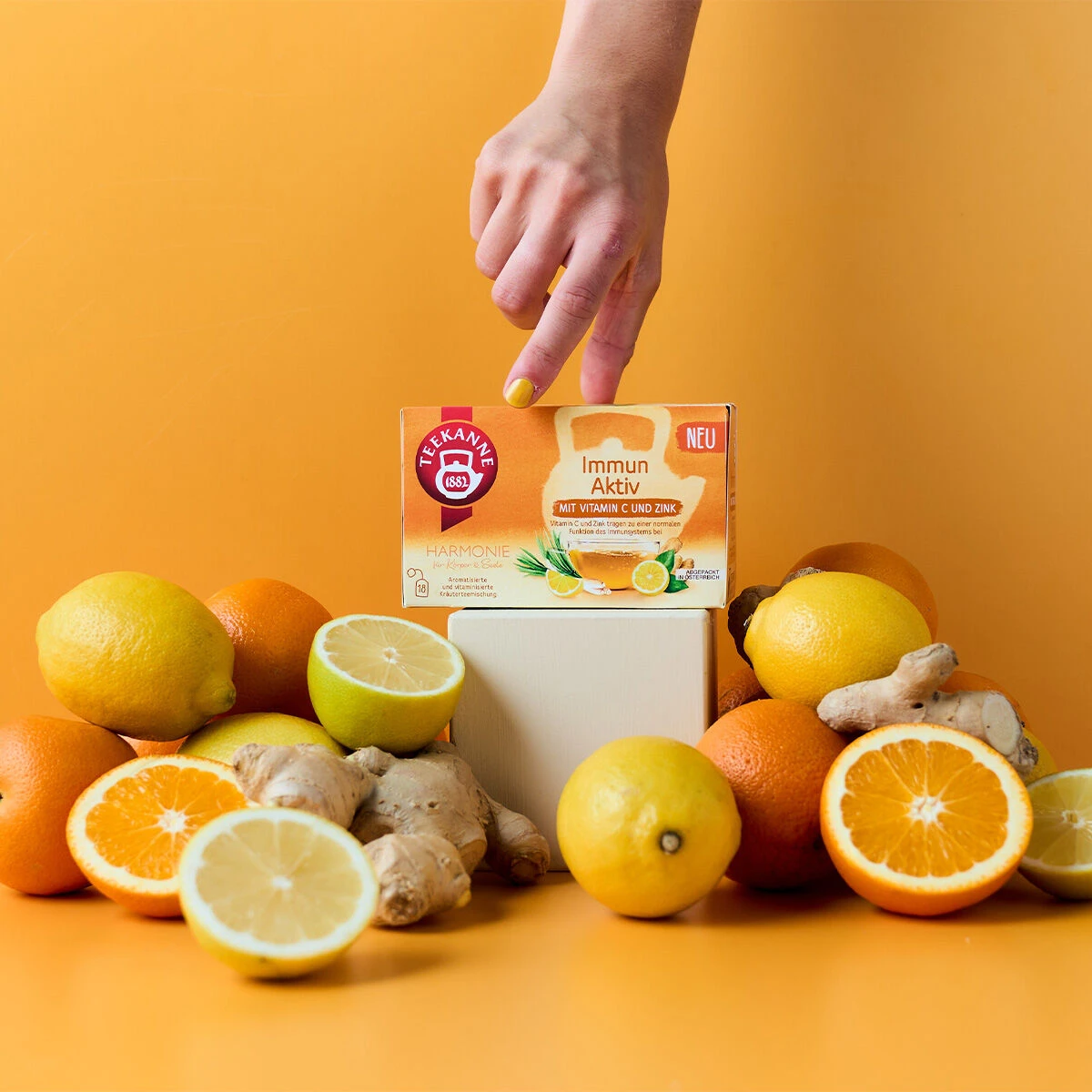 Eine Hand mit gelb lackierten Nägeln zeigt auf eine Schachtel Immun Aktiv Tee, umgeben von Zitronen, Orangen und Ingwer auf gelbem Hintergrund.