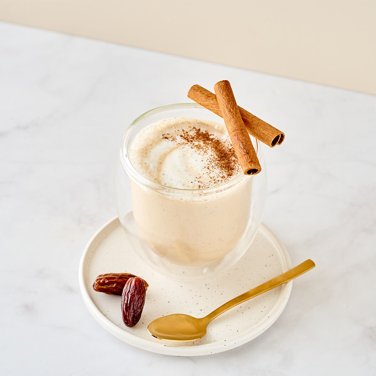 Ein cremiger Indischer Chai-Smoothie in einem Glas, garniert mit Zimtpulver und zwei Zimtstangen, serviert auf einem weißen Teller mit einem goldenen Löffel und einer Dattel, auf einer weißen Marmoroberfläche.