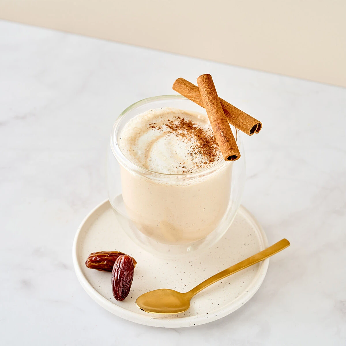 Ein cremiger Indischer Chai-Smoothie in einem Glas, garniert mit Zimtpulver und zwei Zimtstangen, serviert auf einem weißen Teller mit einem goldenen Löffel und einer Dattel, auf einer weißen Marmoroberfläche.