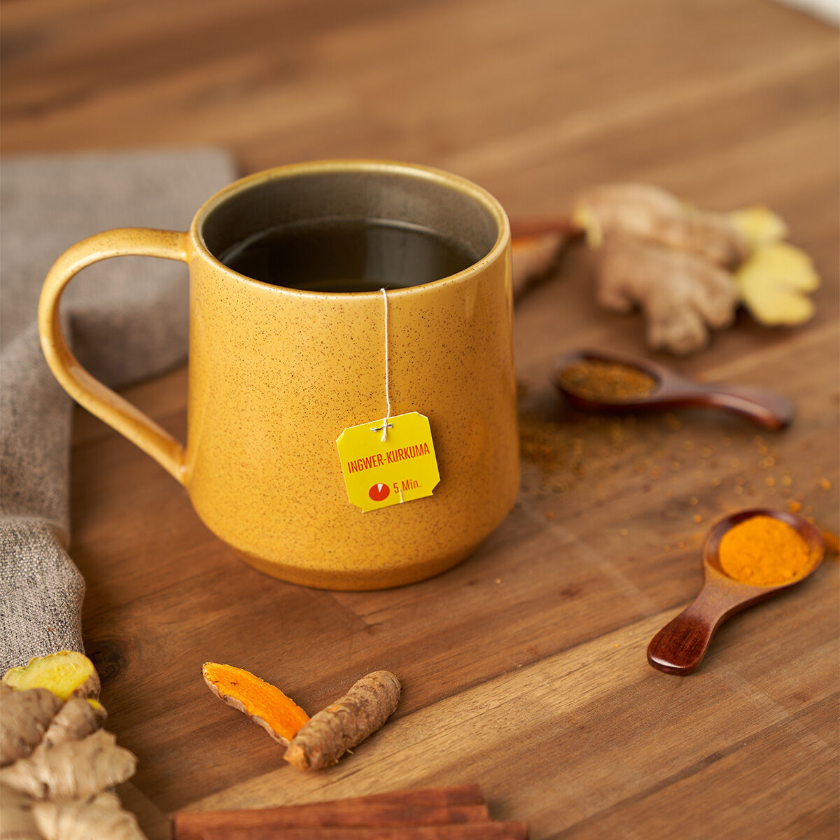 Eine gelbe Tasse Tee mit einem Teebeutel mit der Aufschrift „Ingwer-Kurkuma BIO“ steht auf einem Holztisch, umgeben von Kurkuma-Wurzeln, Ingwer, Zimtstangen, Kräutern und Holzlöffeln voller gemahlener Gewürze.