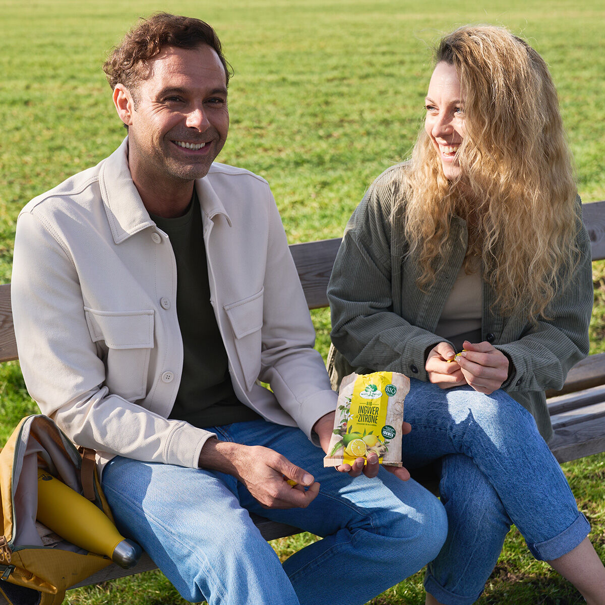 Ein Mann und eine Frau entspannen sich lächelnd auf einer sonnigen Parkbank, während der Mann eine gelbe Packung 65-g-Bonbons mit Ingwer und Zitrone hält, im Hintergrund ist grünes Gras zu sehen.